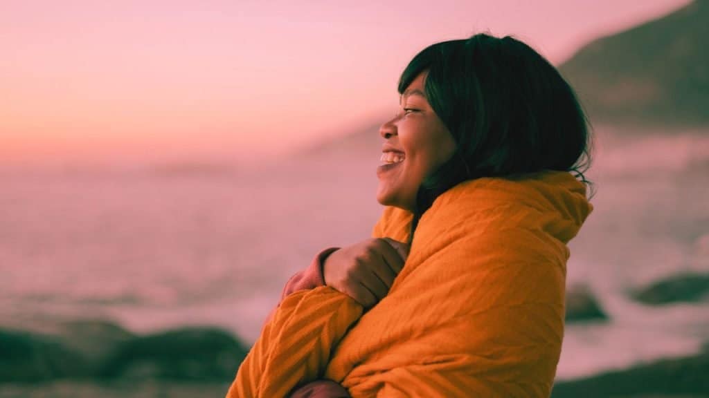 A sober woman wraps herself in a blanket and smiles out at the sea enjoying her newfound emotional sobriety