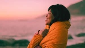 A sober woman wraps herself in a blanket and smiles out at the sea enjoying her newfound emotional sobriety