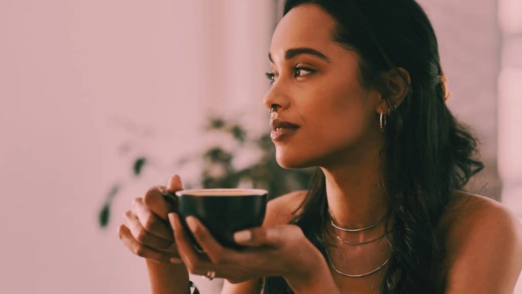 A beautiful woman holds a cup of tea and looks out the window with a serene look on her face