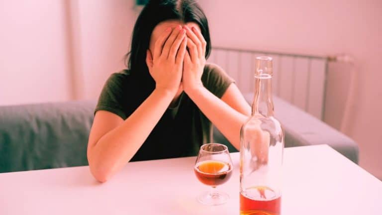 A woman struggling with alcohol cravings covers her face with her hands. There is a wine bottle and wine glass in front of her on the table