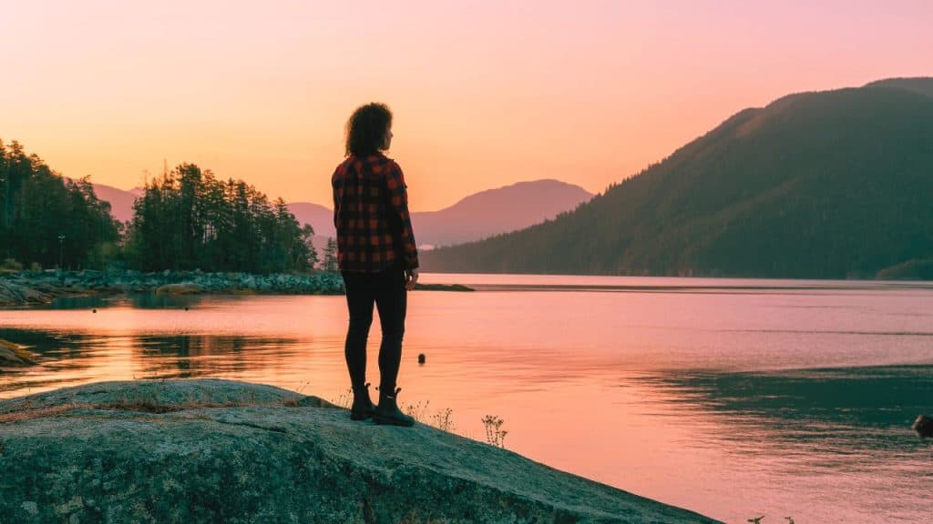 A sober woman staring out at a lake with her back to the camera