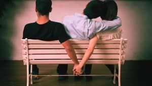 Three people sit on a bench. The woman secretly holds hand with the second man indicating she is about monkey branch and cheat on her current partner beside her