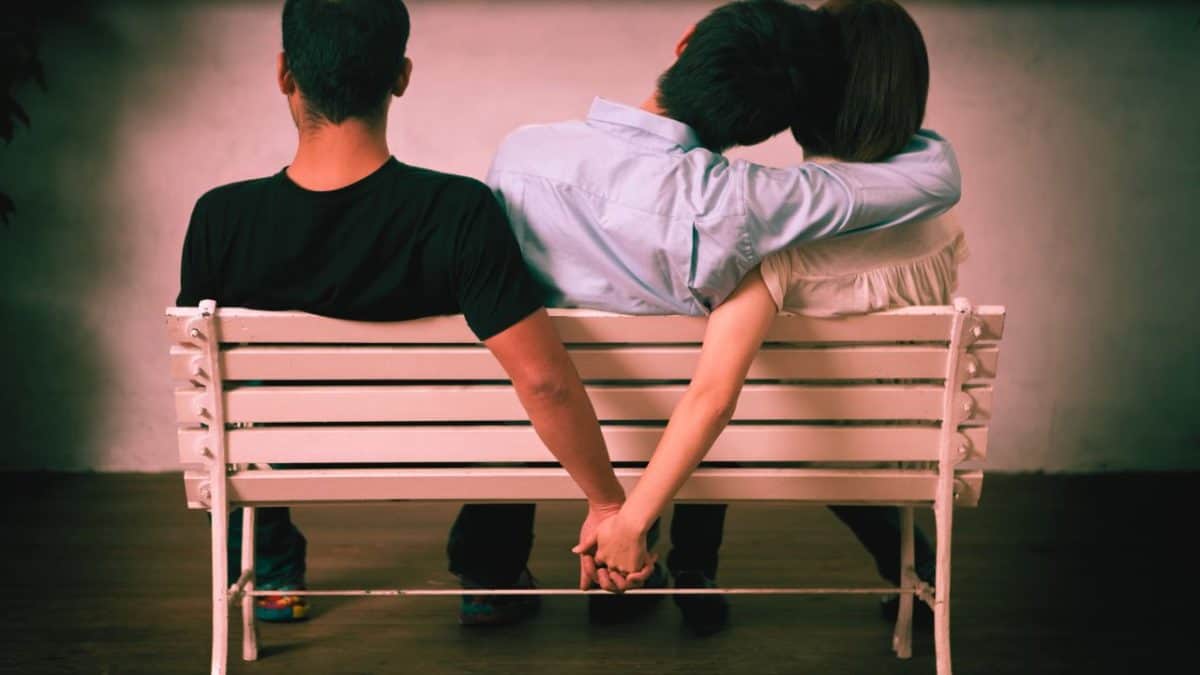 Three people sit on a bench. The woman secretly holds hand with the second man indicating she is about monkey branch and cheat on her current partner beside her