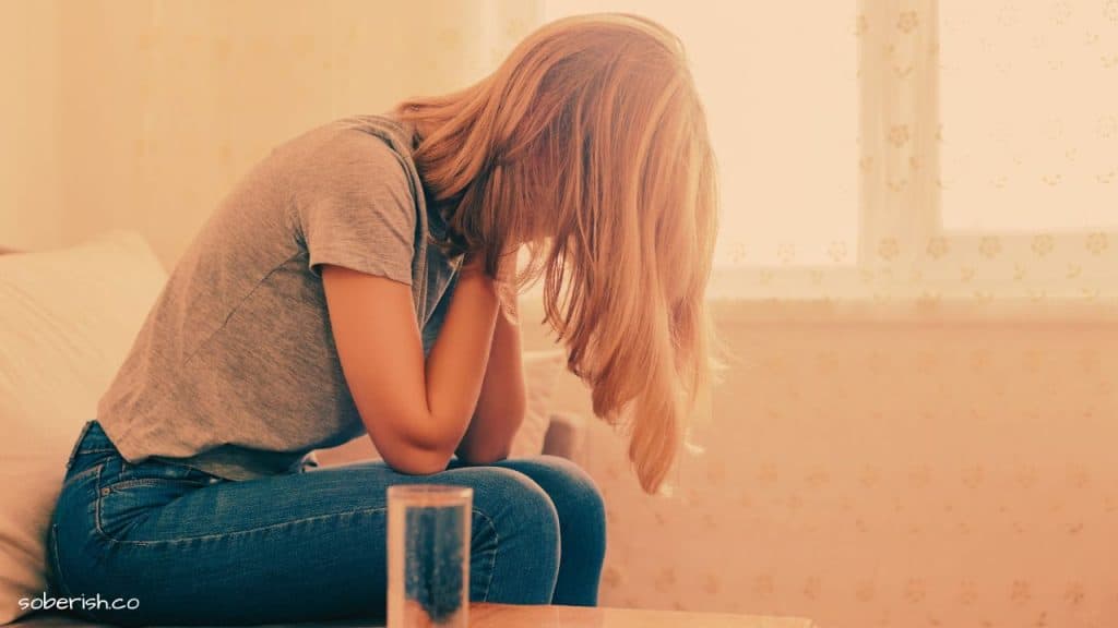 A woman sits on a couch with head in hands, suffering from an alcohol-related headache. There is a glass of water on the table