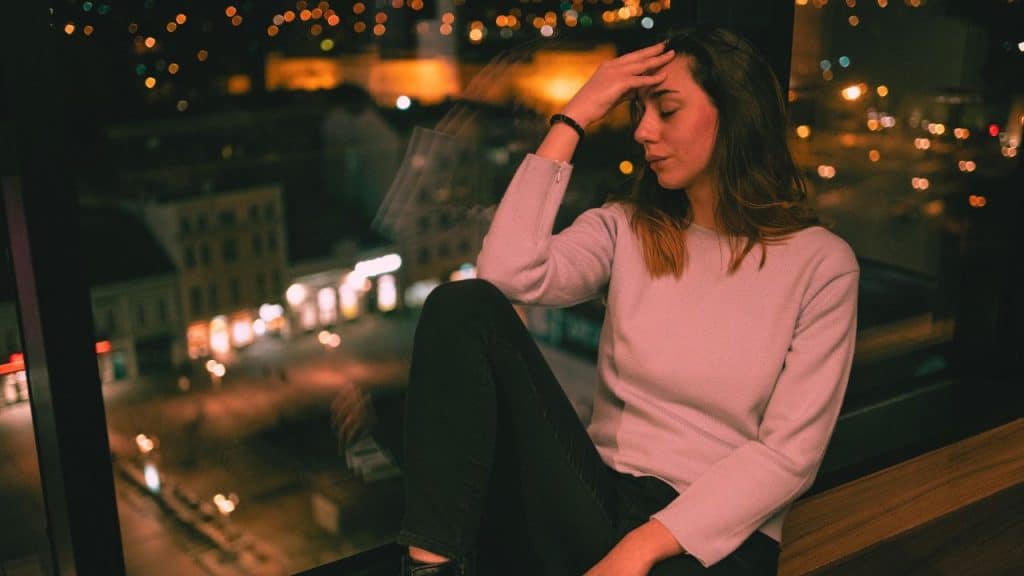 A woman struggling with emotional permanence sits by the ledge of her window stressed, with head in hand