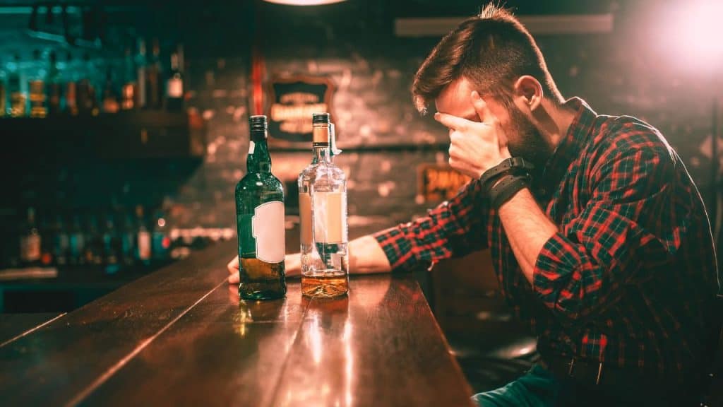 A man sits at a bar angry at himself. There are two mostly drank bottles of whiskey in front of him. He can't seem to stop drinking once he starts