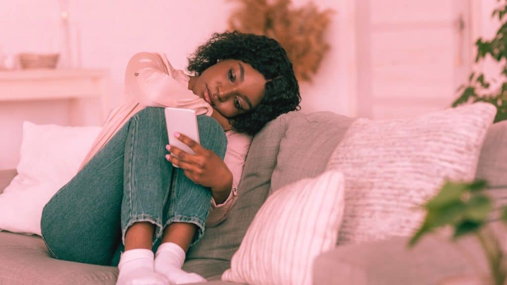A codependent woman sits on the couch staring at her phone, waiting for a text from her partner