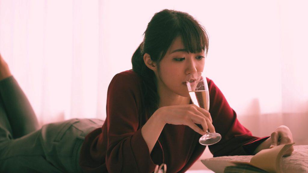 A woman is a high functioning alcoholic. She drinks wine while reading a book.
