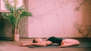A woman in a black leotard lays on a mat doing a somatic exercise for hips with arms back stretching away from the body