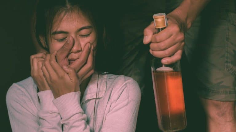 A woman grips her partner's hand that he has clutched around her face. In the other hand he holds a bottle of alcohol. He's an angry drunk.